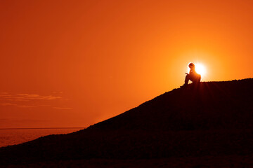 one alone silhouette of relaxing resting woman female girl sitting under sunset light on the sea coast and colorful yellow sky and looking at screen of smartphone. Batumi, Georgia. Black Sea. sunlight