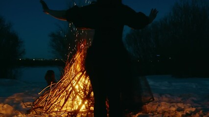 Witch dancing and performing magic ritual near bonfire in winter snowy forest at night, sparks flying around, dark silhouette against firelight and twilight sky, magical atmosphere