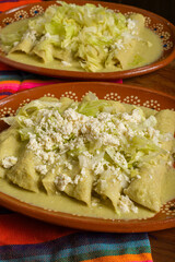 Green enchiladas with lettuce and cheese served in a clay dish. Colorful tablecloth. Close up.