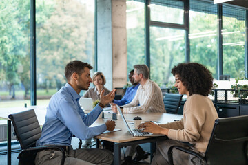 Two business people using laptop on desk discussing strategy company with other employees on background