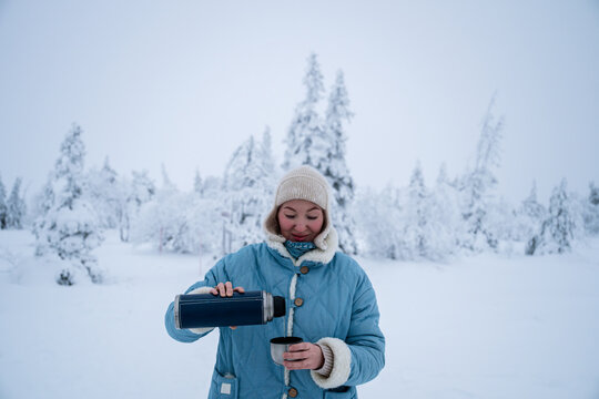 Woman pouring a hot drink from a thermos into a cup in winter forest