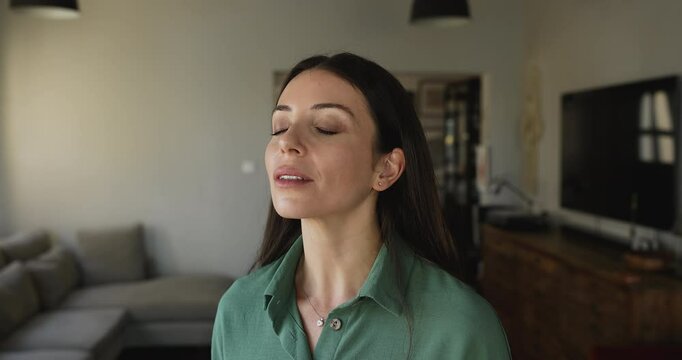 Close up beautiful young woman stand alone in contemporary living room with her eyes closed, breath fresh conditioner air, smile, enjoy freshness inside during summer day. Climate control, stress-free