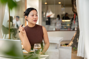 A thoughtful Asian woman looks away, pondering something while working remotely from a cafe.