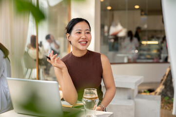 A smiling Asian woman looks away from the camera, daydreaming while working remotely from a cafe.