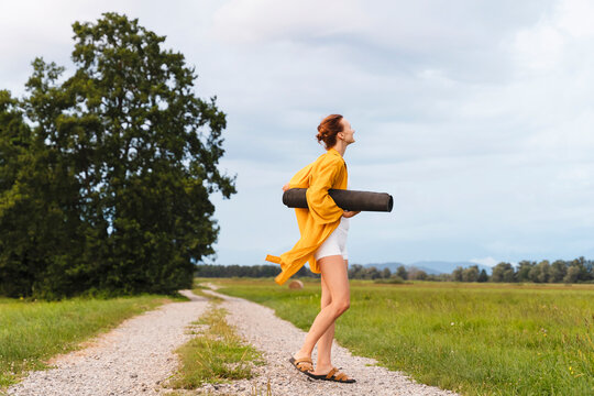Woman holding yoga mat and standing on dust road in nature