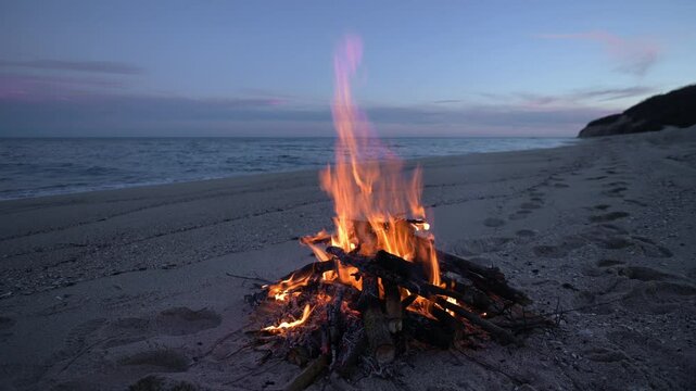 Warm flames of a campfire illuminate a peaceful sandy beach with a calm sea and soft twilight in the background. Ideal for travel, adventure, and serenity concepts
