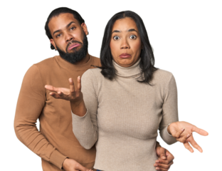Young Latino couple in studio doubting and shrugging shoulders in questioning gesture.