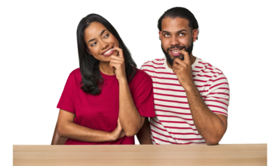Seated young Latino couple at table relaxed thinking about something looking at a copy space.