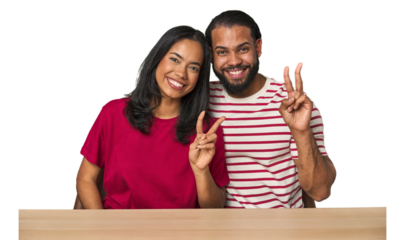 Seated young Latino couple at table showing victory sign and smiling broadly.