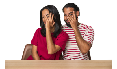Seated young Latino couple at table blink at the camera through fingers, embarrassed covering face.