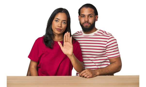 Seated young Latino couple at table standing with outstretched hand showing stop sign, preventing you. - Powered by Adobe