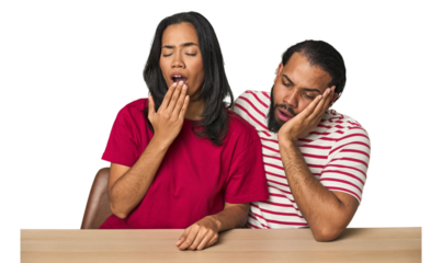 Seated young Latino couple at table yawning showing a tired gesture covering mouth with hand.