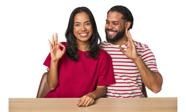 Seated young Latino couple at table cheerful and confident showing ok gesture.