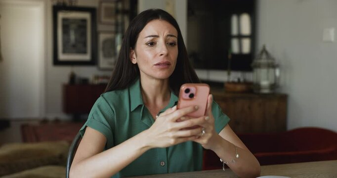 Young woman sit at table at home read news on smartphone, feels dissatisfied, disappointed get bad, awful message in sms looks frustrated, due to on-line scam, missed call, unpaid taxes, bank penalty