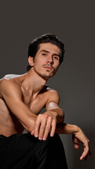 Fototapeta premium Man leaning forward with arms resting on his knees, looking intently into lens. Clean backdrop emphasizes his well-defined features and poised demeanor. Concept of male beauty and facial care,