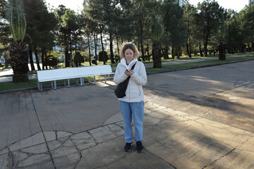 Woman with phone in city park, holding smartphone while walking outside, browsing social media apps.
