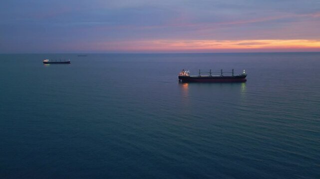 Illuminated cargo ship bulk carrier anchored on the calm sea at twilight, with city lights visible on the horizon
