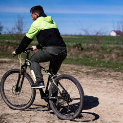 Obraz premium A male cyclist rides a mountain bike on a dirt road. The sun is shining, and the landscape is rural. The rider is wearing a jacket.