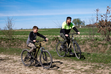 Obraz premium A man and boy cycle along a dirt path, enjoying a sunny day. The open fields and blue sky create a peaceful scene. They ride mountain bikes.
