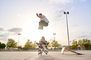 Rollerblader performing a high jump over a seated teammate in a park