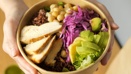 Human hands grasping vibrant salad poki bowl containing grilled chicken, avocado, red cabbage, chickpeas, lettuce, brown rice representing nutritious, wholesome eating