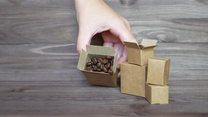 Hand takes small open boxes while coffee beans and empties them on a wooden table, showcasing packaging and coffee products. - Powered by Adobe