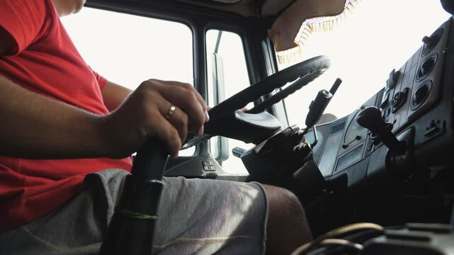 Male hand is shifting gears in a manual transmission while riding a truck. Arm of lorry driver holding a big steering wheel of a car. Trucker driving to destination at country road. Logistics concept