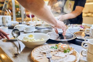 A hand reaches out to take a portion of a variety of dishes from the buffet, surrounded by many people enjoying a meal at a lively event. High quality photo