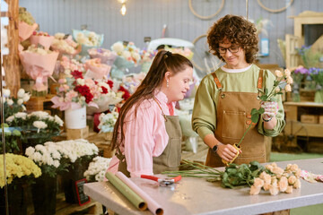 Medium close up of content florist holding rose and demonstrating brand new thorn remover tool to her colleague with down syndrome
