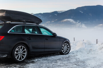Scenic view black modern wagon car with roof rack box parked against foggy morning winter mountain alpine resort nature landscape. Family holidays snowy journey travel auto trip