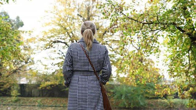 Woman in houndstooth coat standing in public park and enjoying view. Autumn fashion clothing