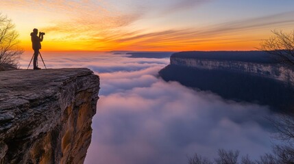 Fototapeta premium Photographer silhouetted on clifftop, capturing sunrise over misty canyon.