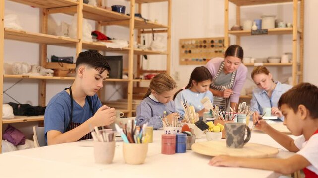 Children and teenagers sit at a table and paint ceramics at the workshop. Teacher shows how to paint a clay product after firing