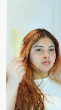 Close up vertical video of freckled woman with red hair in front of mirror combing her hair with wooden eco friendly comb, lifestyle at home