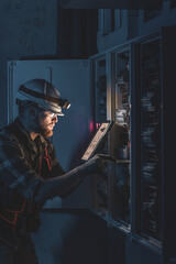 Electrician in overalls, focused on work in switchboard with fuses, using tablet.