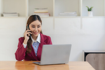 Asian woman uses smartphone while sitting at table with laptop computer at office She writes notes on document graphs. female financial accountant with financial growth statistics chart