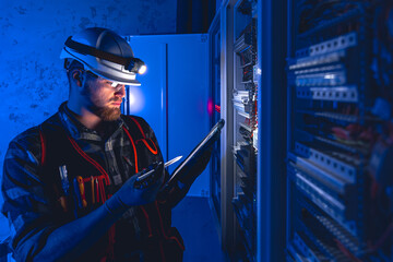 Electrician in overalls, focused on work in switchboard with fuses, using tablet.