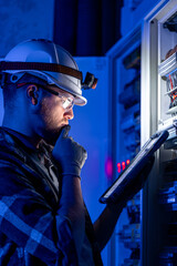 Electrician in overalls, focused on work in switchboard with fuses, using tablet.