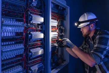 A male electrician works in a switchboard in overalls against the backdrop of emergency lighting.