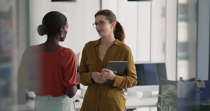 African and Caucasian women colleagues lead discussion standing in contemporary office, smiling, having friendly work-related conversation, shaking hands while finalizing talk. Teamwork, companionship