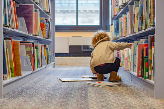 Toddler selecting books from a library shelf with books on the floor. Cleveland, Ohio, USA