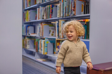 Toddler with curly hair smiling and walking in a library with bookshelves in the background. Cleveland, Ohio, USA