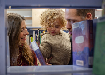A family enjoys time together browsing books at a library, with a toddler in the arms of a smiling man and an engaged woman by their side. Cleveland, Ohio, USA