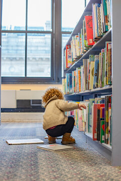 Toddler in a beige sweater and brown boots reaches for a book on a library shelf with large windows in the background. Cleveland, Ohio, USA