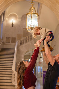 Parents lift their toddler high in an elegant hallway, the child's joy is evident as they reach towards a hanging chandelier. Cleveland Public Library, Ohio, USA
