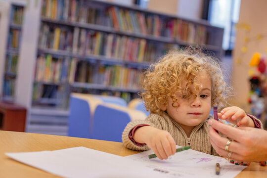 A curly-haired toddler engaging in coloring activity at a table with adult assistance in a library. Cleveland, Ohio, USA