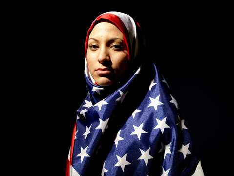 A woman draped in an American flag hijab looks contemplatively to the side against a black background.