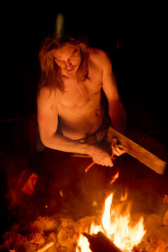 Shirtless man with long hair stoking a campfire at night.