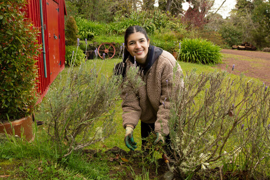 Smiling woman gardening outside wearing gloves near a red shed and greenery, Auckland, New Zealand