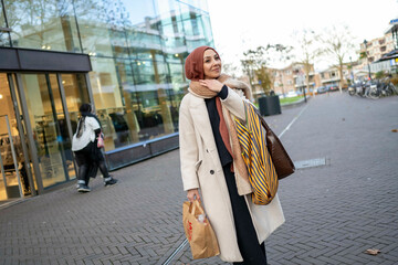 Elegant woman in a hijab and coat smiling while holding shopping bags on a city street, Netherlands
